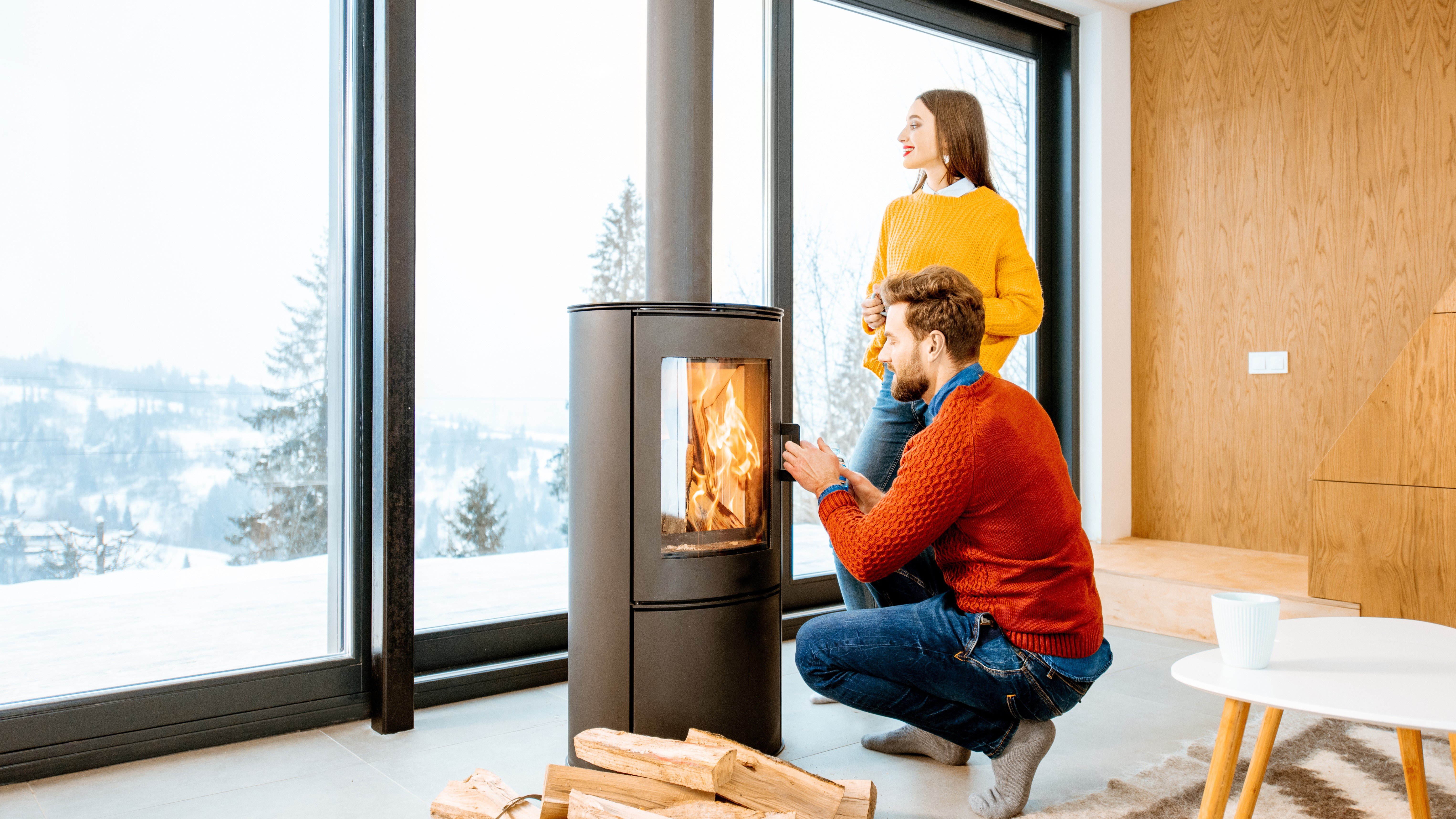A young couple burning firewoods in modern fireplace  front of the large window with wintery landscape.